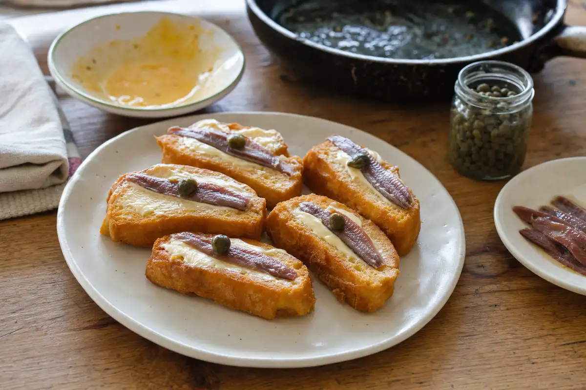 Crostini di Cazzimperio con burro, alici e capperi su piatto di ceramica, con padella e vasetto sul tavolo