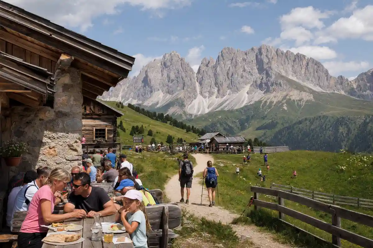 Escursionisti e famiglie a tavola fuori da una baita, con sentiero e cime dolomitiche sullo sfondo