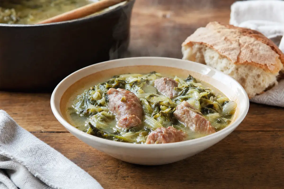 Ciotola di minestra con verdure a foglia e pezzi di carne di maiale su tavolo di legno, con pane rustico