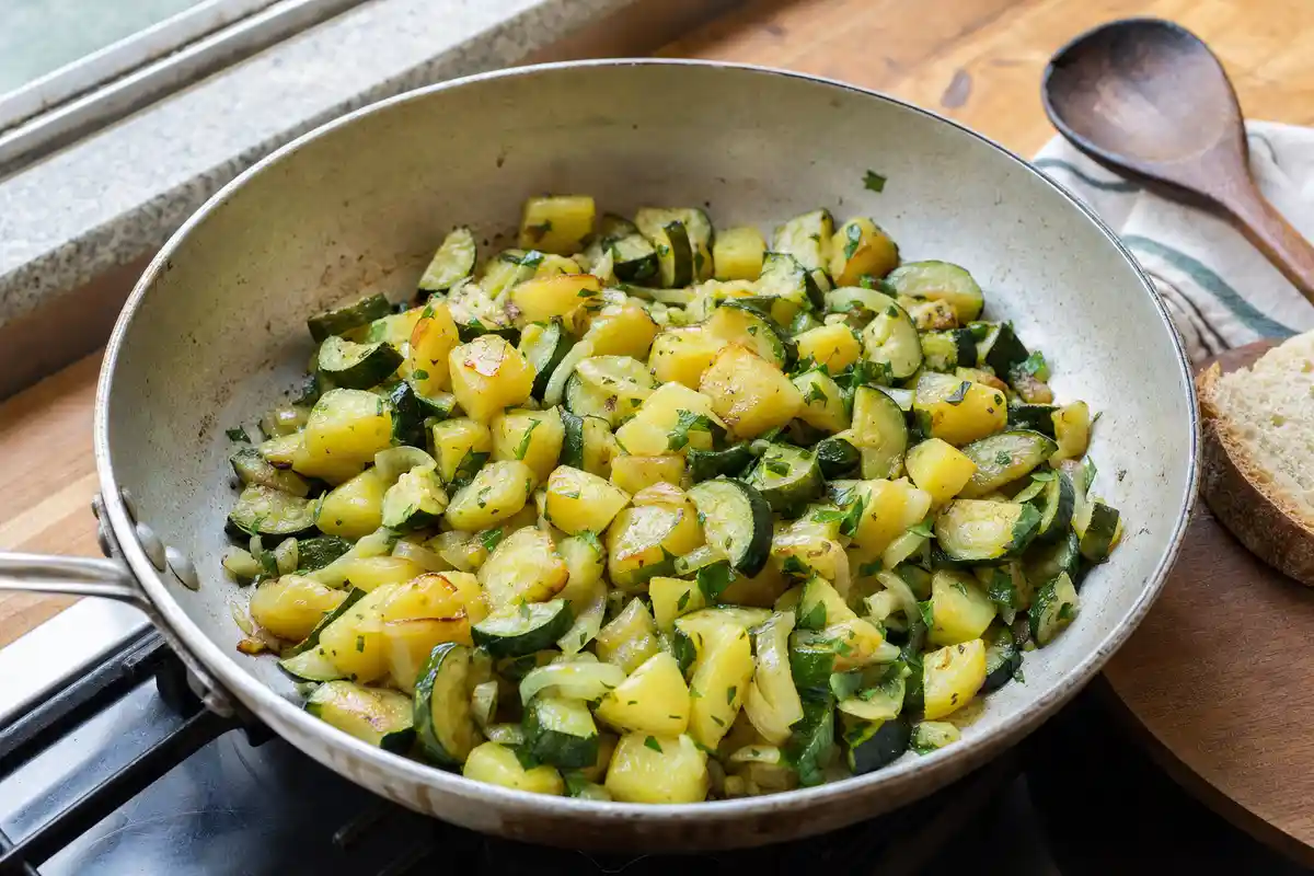 Padella con zucchine e patate a tocchetti con cipolla e prezzemolo, accanto a cucchiaio di legno e pane