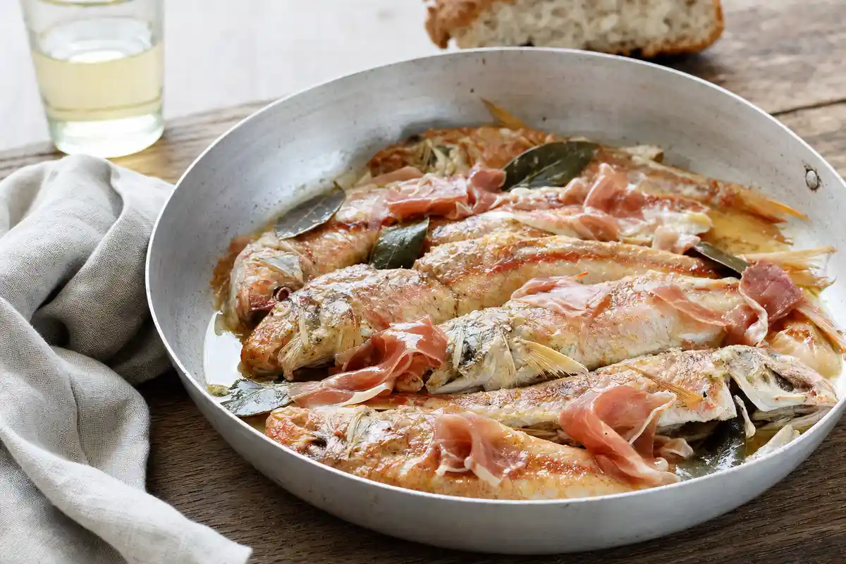 Triglie cotte in teglia con prosciutto crudo e foglie di alloro su tavolo rustico