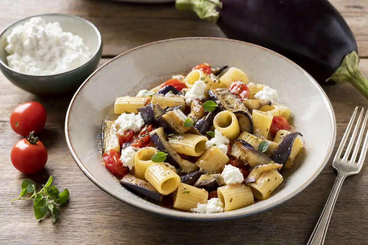 Piatto di mezze maniche con melanzane, pomodorini e ricotta fresca su tavolo di legno, con forchetta a lato