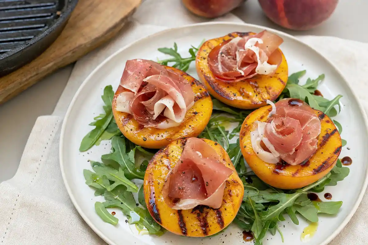 Mezze pesche gialle grigliate con prosciutto crudo su letto di rucola, con olio e gocce di balsamico